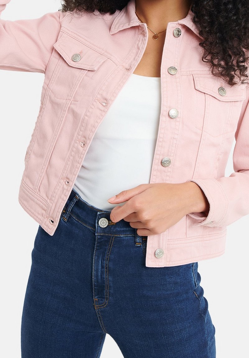Femme portant une veste en denim rose clair sur un haut blanc, attachant un jean bleu foncé à la taille devant un fond uni.