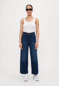 Løse denimjeans i mørkeblå, sammen med en tettsittende hvit tanktop og svarte solbriller. Har høyt liv og frontlukking med knapper.