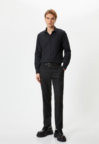 Svart skjorta med knappar, svart bälte och svarta byxor med en glatt yta; modellen har på sig tjocksålade svarta skor. Minimalistisk design.