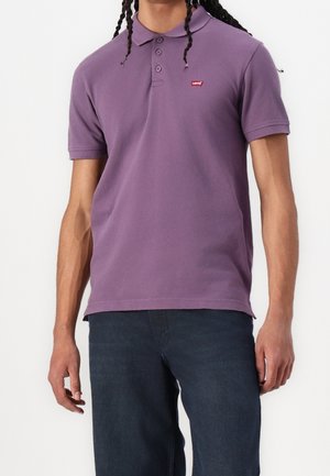 Personne portant un polo violet à manches courtes avec un petit logo rouge Levi's sur la poitrine, associé à un jean bleu foncé.