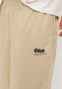 Shorts en mesh beige avec taille élastique et logo texte noir indiquant « Shine » et texte plus petit « Ceux qui suivent la lumière finiront par briller intensément. »