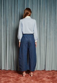 Mulher em frente a cortinas azuis, vestindo uma blusa azul clara, calças largas azul-marinho brilhantes e sandálias de salto alto bege.