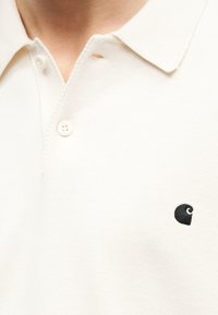 Κοντινό πλάνο ενός ατόμου που φοράει ένα μπεζ πουκάμισο πόλο με δύο κουμπιά και ένα μικρό μαύρο κεντητό λογότυπο στο στήθος.