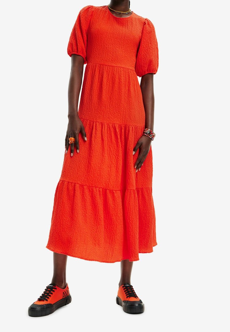 Personne en robe midi texturée orange vif avec manches bouffantes, portant des baskets orange et noires assorties et des bracelets et bagues colorés.