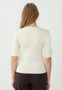 Beige aansluitend topje met korte mouwen, gladde textuur en ronde halslijn. G gedragen met donkerbruine broek, met een eenvoudig ontwerp aan de achterkant.