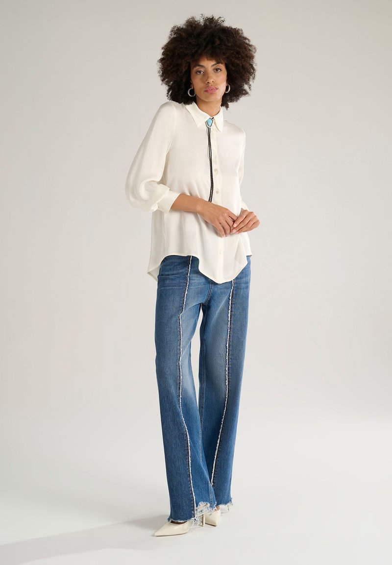 Blusa bianca a maniche lunghe con colletto, dotata di un gancetto nero e un accento turchese, abbinata a jeans blu con cuciture a contrasto e orlo sfrangiato.