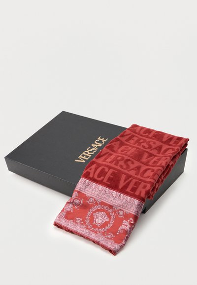 Κόκκινη πετσέτα Versace με το μοτίβο της Μέδουσας, διπλωμένη πάνω σε μαύρο κουτί Versace με χρυσό λογότυπο σε λευκή επιφάνεια.