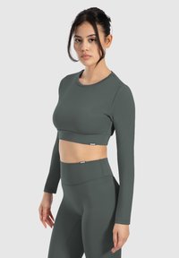 Dunkelgrünes, langärmliges Cropped-Top mit einer figurbetonten Passform. Glatte Textur und runder Halsausschnitt. Kombiniert mit hochgeschnittenen, passenden Leggings.