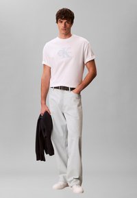 Άνδρας που φοράει λευκό t-shirt Calvin Klein και ανοιχτόχρωμο τζιν, κρατώντας μαύρο σακάκι στο αριστερό χέρι, στέκεται μπροστά σε γκρι φόντο.