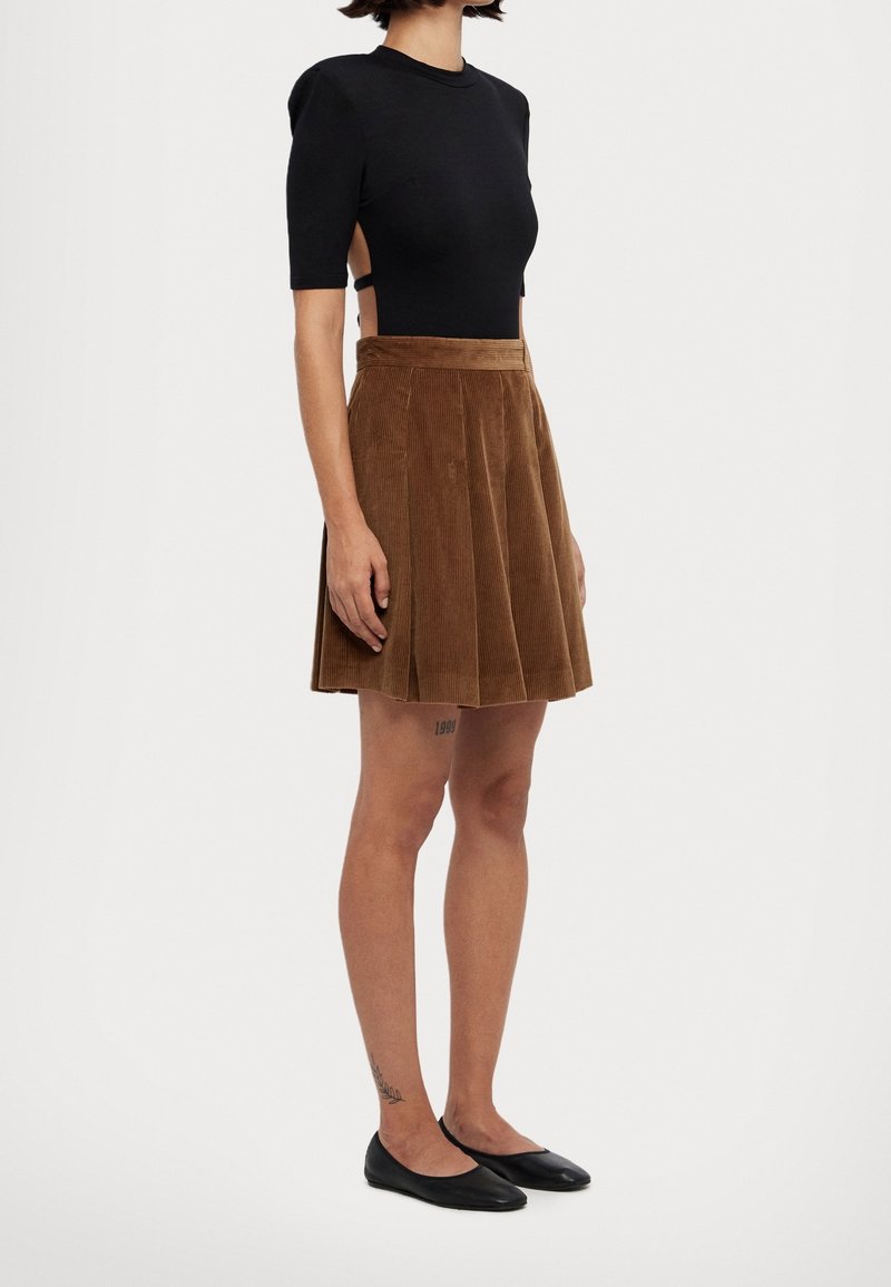 Femme portant un haut noir à manches courtes avec découpes latérales, jupe plissée en velours côtelé marron, ballerines noires et tatouages sur la jambe et la cheville.