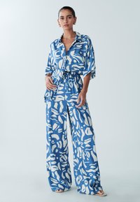 Ensemble chemise et pantalon à motifs bleus et blancs ; coupe ample, manches courtes, fermeture à boutons, taille élastique, formes abstraites sur le tissu.