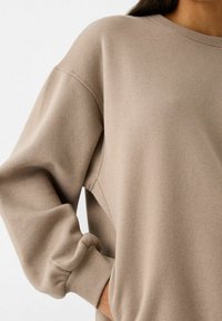 Mujer vistiendo un suéter beige de manga larga con puños acanalados, mano parcialmente en el bolsillo, y cabello oscuro visible sobre un fondo blanco.