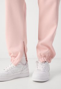 Ανοιχτό ροζ φόρμες με φερμουάρ στους αστραγάλους και συγκεντρωμένα μανίκια, συνδυασμένα με λευκά αθλητικά παπούτσια με σχέδιο, σε ουδέτερο φόντο.