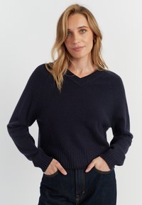 Pull en maille bleu marine avec un col en V, des poignets côtelés et un ourlet côtelé. Texture douce avec une coupe décontractée, associé à un jean noir pour le contraste.