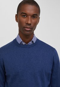 Maglione azzurro lavorato a maglia con colletto rotondo, indossato sopra una camicia colletto a righe bianche e blu. Il maglione presenta una texture morbida e una silhouette aderente.
