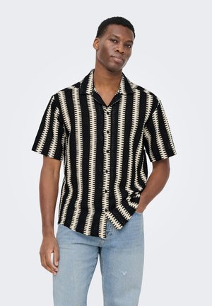 Mężczyzna stojący z ręką w kieszeni, ubrany w czarno-białą koszulę z krótkim rękawem w pionowe paski oraz jasnoniebieskie dżinsy na jednolitym tle.