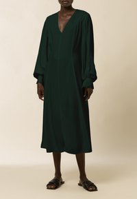 Vestido verde oscuro de manga larga con escote en V profundo y un corte relajado. Tejido suave, de longitud media, con mangas fruncidas y costuras mínimas.