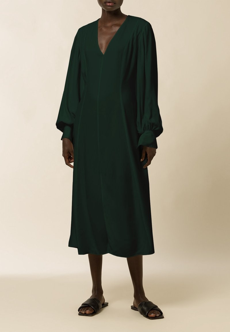 Vestido verde oscuro de manga larga con escote en V profundo y un corte relajado. Tejido suave, de longitud media, con mangas fruncidas y costuras mínimas.
