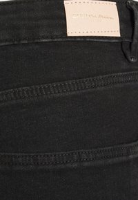 Tecido de ganga preto com uma textura suave, apresentando costuras contrastantes e um rótulo de pele castanha com o nome da marca em relevo na cinta.