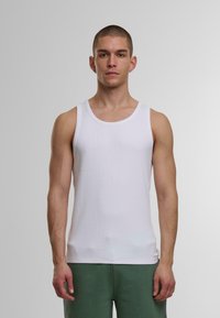 Helles Tanktop aus geripptem Baumwollstoff, mit Rundhalsausschnitt und weiten Armöffnungen, getragen mit grünen Jogginghosen.