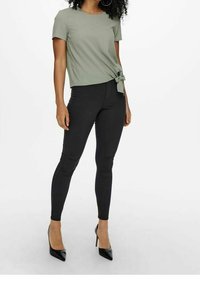 Top verde oliva a maniche corte con dettaglio nodo, abbinato a pantaloni skinny neri e décolleté neri a punta. Tessuto morbido e design aderente.