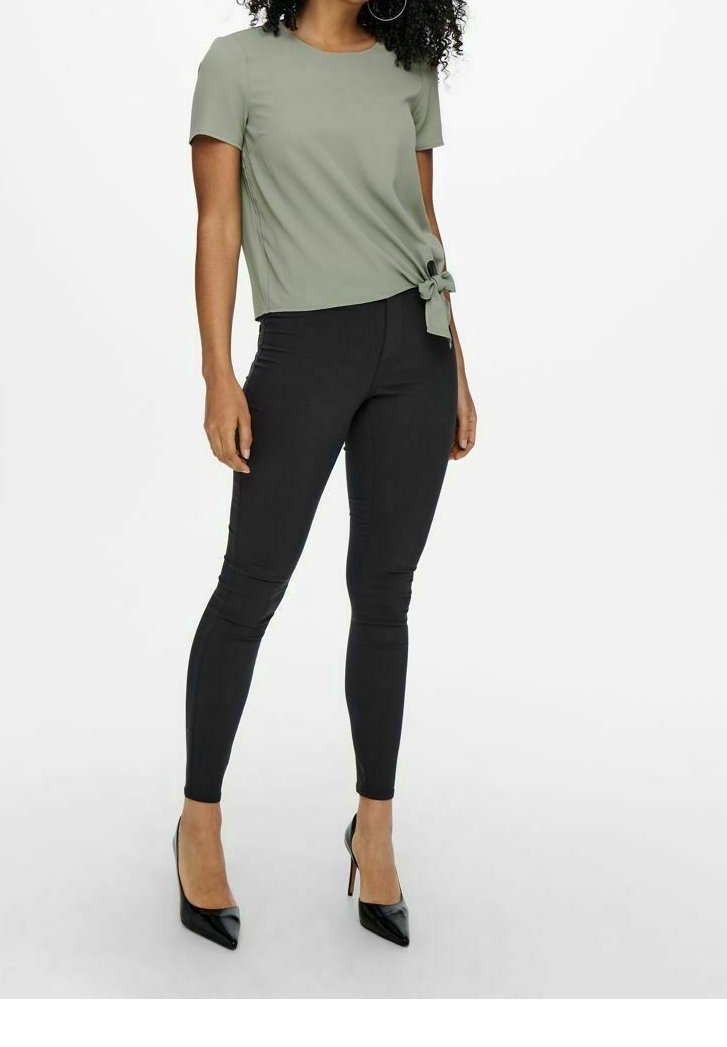 Top verde oliva a maniche corte con dettaglio nodo, abbinato a pantaloni skinny neri e décolleté neri a punta. Tessuto morbido e design aderente.