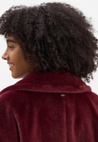 Vrouw met krullend haar die glimlacht, gekleed in een zachte, diep rode jas met een brede kraag, van opzij en van achteren gezien.