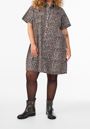 Kobieta ubrana w sukienkę w panterkę z krótkim rękawem i zapinana na guziki, czarne rajstopy oraz ciemne botki na kostkę na białym tle.
