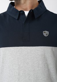 Μπλε ναυτικό και γκρι πόλο από βαμβάκι, με γιακά, δύο κουμπιά και λογότυπο ασπίδας στο αριστερό στήθος.