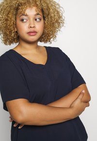 Navy blue v-neck top with short sleeves, made of lightweight fabric. Features a subtle textured surface and relaxed fit.