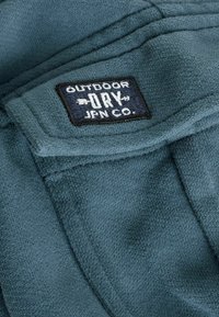 Tessuto in pile blu con una toppa rettangolare cucita che riporta la scritta "OUTDOOR DRY JPN CO." in lettere bianche su uno sfondo blu navy.
