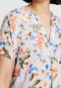 Blomstret bluse i blødt pink stof med mønstre af blå, orange og grønne blomster. Den har en V-hals og korte ærmer med en tekstureret overflade.