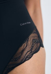 Cuecas pretas Calvin Klein com acabamento em renda usadas na anca de uma pessoa contra um fundo neutro.