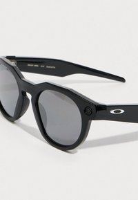 Mustat oktaaliset aurinkolasit tummilla linsseillä, joissa on kiiltävä pinta, sisäänrakennettu kamera ja hopeinen logo aksentti varressa.