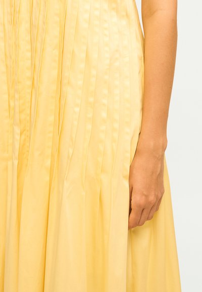 Κοντινή λήψη ενός κίτρινου πλισέ φουστακιού με το δεξί χέρι ενός ατόμου να στηρίζεται δίπλα του σε απλό φόντο.