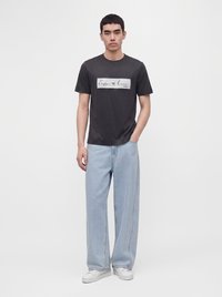 Мъж, носещ черна тениска Emporio Armani, светлосини дънки с широки крачоли и бели маратонки, стои с една ръка в джоба си.