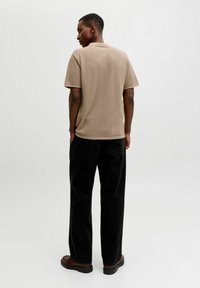 Un homme debout de dos portant un polo en maille beige à manches courtes, un pantalon noir ample, et des chaussures marron sur un fond clair uni.