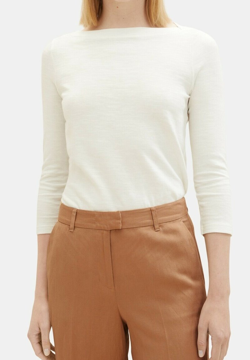 Kobieta w dopasowanej białej bluzce z długim rękawem, włożonej w brązowe spodnie o prostym kroju i wysokim stanie, z pętelkami na pasek.