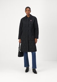 Abrigo largo negro acolchado con cuello, que cuenta con botones delanteros, bolsillos laterales y un pequeño logo rojo y blanco. Combinado con pantalones vaqueros acampanados azules.