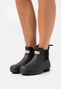 Bottes de cheville en caoutchouc noir avec une semelle texturée, des panneaux latéraux élastiques et un logo "HUNTER" rouge et blanc bien en vue à l'avant.