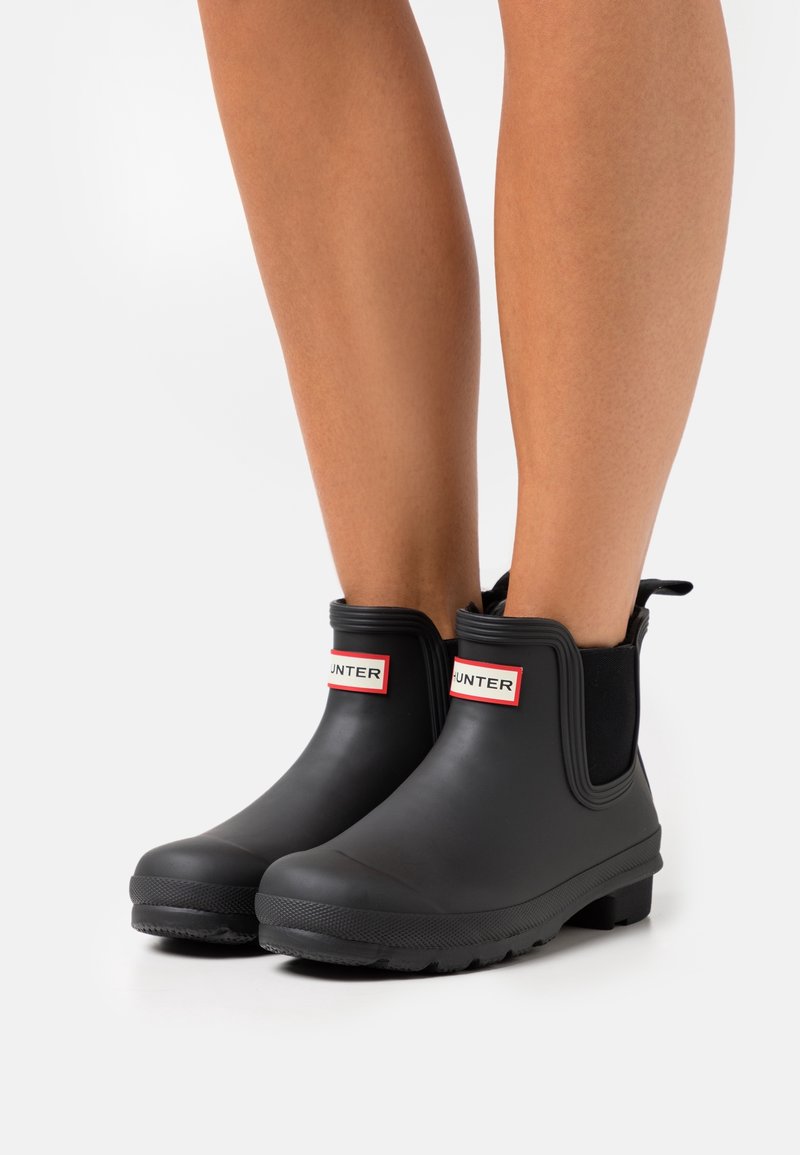Bottes de cheville en caoutchouc noir avec une semelle texturée, des panneaux latéraux élastiques et un logo "HUNTER" rouge et blanc bien en vue à l'avant.