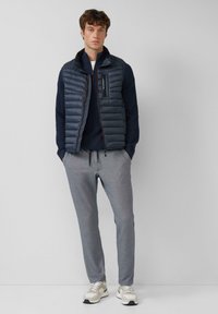 Marineblauwe puffer vest over een marineblauwe gebreide top, gecombineerd met grijze sweatpants. Heeft een ritszak op de borst. Schoenen zijn lichtgrijs met donkere accenten.