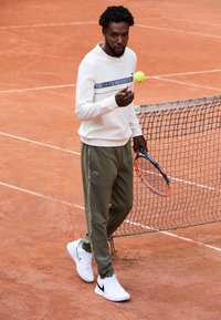 Mann i hvit sweatshirt og grønne treningsbukser holder en tenniskølle og hopper med en tennisball på leircourt nær nettet.