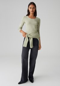 Hellgrünes, enganliegendes Langarmoberteil, kombiniert mit hochgeschnittenen dunklen Jeans. Ein weicher, grüner Strickpullover, der an der Taille gebunden ist, fügt Textur hinzu.