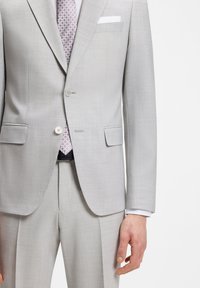 Ανοιχτό γκρι κοστούμι προσαρμοσμένης γραμμής με σακάκι δύο κουμπιών, ταιριαστό παντελόνι, λευκό πουκάμισο και γραβάτα με σχέδιο σε άτομο που στέκεται μπροστά σε λευκό φόντο.