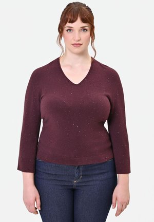 Donna con pelle chiara e capelli rossi che indossa un maglione bordeaux a scollo a V con brillantini e jeans blu scuro, in piedi davanti a uno sfondo chiaro e semplice.
