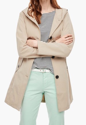 Veste imperméable beige avec capuche, boutons à l'avant et deux poches, portée sur un haut rayé et un pantalon vert menthe.