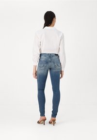 Jean bleu ajusté avec une légère décoloration, présentant une taille haute et des poches arrière. Associé à une chemise blanche à col et des talons marron clair.