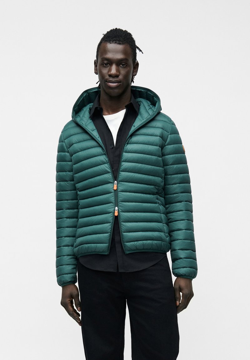 Άνδρας που φοράει πράσινο-μπλε καπιτονέ μπουφάν με κουκούλα πάνω από μαύρο πουκάμισο και λευκό εσώρουχο, στέκεται μπροστά σε απλό ανοιχτό φόντο.