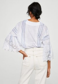 Blouse en lin blanc à rayures verticales bleues, manches évasées en dentelle avec motif ajouré, associée à un pantalon taille haute écru.